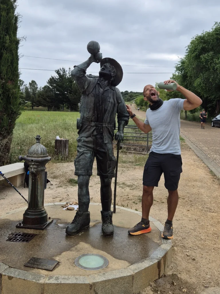 Simon pretending to drink water next to a bronze statue on the Camino de Santiago
